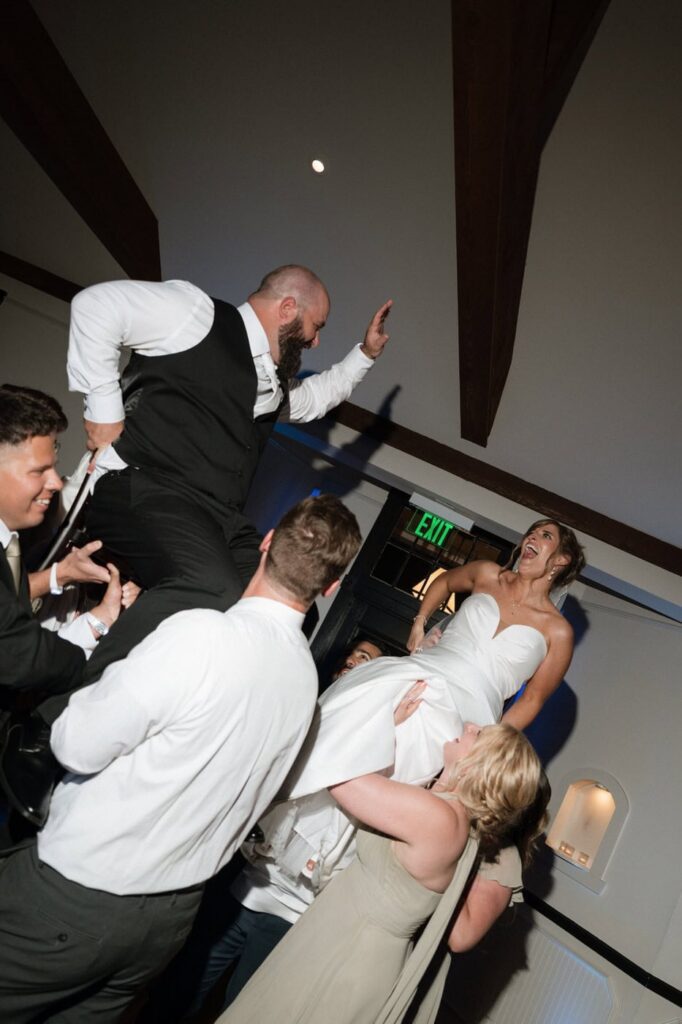bride and groom laughing while being lifted in chairs at their wedding reception